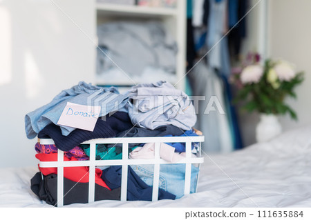 A basket of selected clothes for donating to a Charity shop standing on the bed. Decluttering Clothes, Sorting, And Cleaning Up. Reuse, second-hand. Conscious consumer, sustainability. Selective focus A basket of selected clothes for donating to a Charity shop standing on the bed. Decluttering Clothes, Sorting, And Cleaning Up. Reuse, second-hand. Conscious consumer, sustainability. Selective focus 111635884