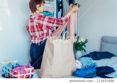 Woman selecting clothes from her wardrobe for donating to a Charity shop. Decluttering, Sorting clothes and Cleaning Up. Reuse, second-hand concept. Conscious consumer, sustainable lifestyle. 111635886