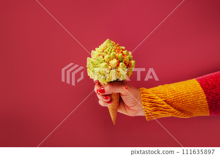 Female hand holding Waffle cone with Romanesco cabbage with dripping fruit sauce as vegetarian dessert on bright red background. Plant based Ice cream. Creative healthy vegetarian and vegan food. Female hand holding Waffle cone with Romanesco cabbage with dripping fruit sauce as vegetarian dessert on bright red background. Plant based Ice cream. Creative healthy vegetarian and vegan food. 111635897