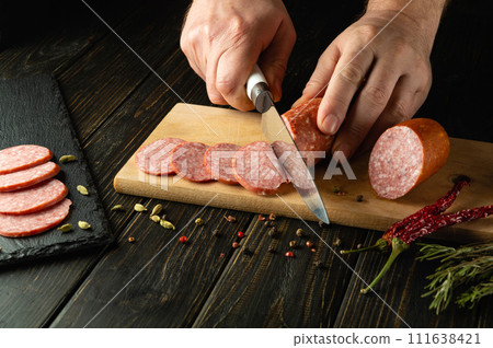 Slicing salami sausage with a knife in the hand of a chef for serving a festive table in a restaurant Slicing salami sausage with a knife in the hand of a chef for serving a festive table in a restaurant 111638421