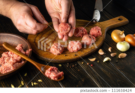 The chef hand holds a ground beef meatball ready for cooking. The concept of cooking cutlets on the kitchen table with aromatic spices 111638424
