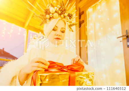 A woman in a white dress is holding a gold box with a red ribbon. She is wearing a crown on her head. The woman appears to be opening the gift box. 111640312