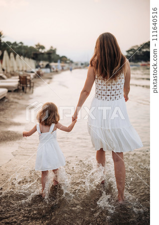 A little girl and a young mother in white dresses walk barefoot on the sand by the sea at sunset.  111640516