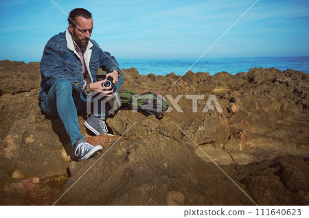 Caucasian bearded hiker adventurer sitting on the rocky cliff by seam holding a stainless steel thermos with hot drink Caucasian bearded hiker adventurer sitting on the rocky cliff by seam holding a stainless steel thermos with hot drink 111640623