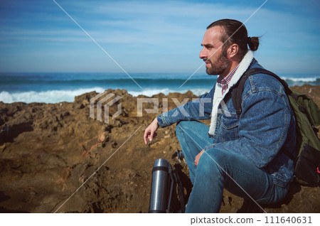 Bearded serious male traveler sitting on the rock by sea, enjoying his coffee break outdoor Bearded serious male traveler sitting on the rock by sea, enjoying his coffee break outdoor 111640631