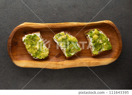 toast with cream cheese and avocado on a wooden plate, dark background 111643395