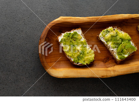 toast with cream cheese and avocado on a wooden plate, dark background 111643400