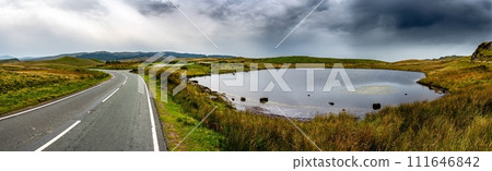 Narrow Road Beneath Small Pond In Wild Landscape Of Snowdonia National Park in Wales, United Kingdom 111646842