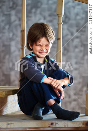 Dissatisfied grimace of little boy, 7 years old, sitting on step of wooden staircase leading to second floor of house in suburbs. Dissatisfied grimace of little boy, 7 years old, sitting on step of wooden staircase leading to second floor of house in suburbs. 111647557