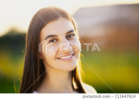 Portrait of white brunette woman in her 20s , in light of setting sun against blurred background of rural landscape, close-up. Portrait of white brunette woman in her 20s , in light of setting sun against blurred background of rural landscape, close-up. 111647624