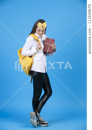 Confident young girl wearing funny headband smiling, while carrying yellow backpack indoors. Side view of joyful female student holding books, while looking down, isolated on blue. Concept of posing. 111648678