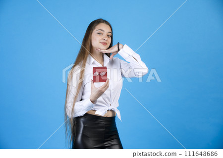 Pretty girl with long hair demonstrating small gift box, while posing at camera. Cute lady with lovely present, putting chin on hand, while smiling at camera, isolated on blue. Concept of posing. 111648686