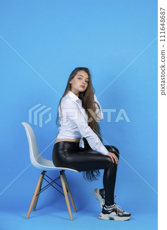 Pensive, graceful lady in modern outfit sexually posing, while sitting in cozy chair. Side view of gorgeous girl touching loose fair hair, while looking at camera, isolated on blue. Concept f posing. 111648687