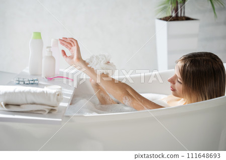 Happy caucasian lady washing arm with soapy sponge in bath. Side view of pretty young woman lather body with washcloth, while soaking in bathtub, with copy space. Concept of bathing, hygiene. Happy caucasian lady washing arm with soapy sponge in bath. Side view of pretty young woman lather body with washcloth, while soaking in bathtub, with copy space. Concept of bathing, hygiene. 111648693