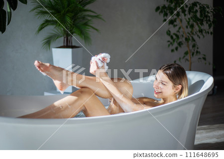 Smiling blonde lady washing legs with soapy sponge in bath in morning. Side view of beautiful caucasian woman lather body with washcloth, while soaking in bathtub. Concept of relax, bathing process. 111648695