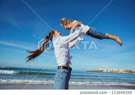 Happy mother holding her little daughter girl in her outstretched hands, throwing up and playing on the sandy beach Happy mother holding her little daughter girl in her outstretched hands, throwing up and playing on the sandy beach 111649169
