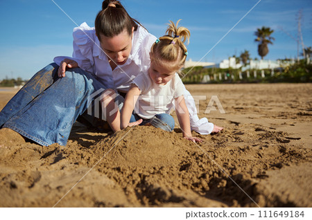Happy woman and her cute little child girl enjoying time together, sitting on the sandy beach on warm sunny day Happy woman and her cute little child girl enjoying time together, sitting on the sandy beach on warm sunny day 111649184