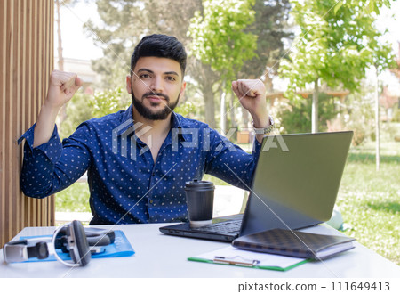Front view of brunette asain guy working at laptop outdoors. Handsome young businessman looking at camera, raising hands, showing hurray. Concept of urban lifestyle and youth. 111649413