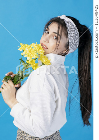 Side view of young, brunette woman with hair band posing with yellow chrysanthemum, looking at camera. Concept of beauty. Side view of young, brunette woman with hair band posing with yellow chrysanthemum, looking at camera. Concept of beauty. 111649425
