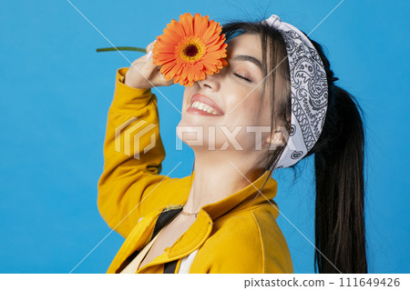 Side view of pretty, young woman posing with orange flower, raising hand, touching face, smiling. Isolated on blue studio background. Side view of pretty, young woman posing with orange flower, raising hand, touching face, smiling. Isolated on blue studio background. 111649426