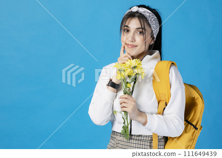 Front view of pretty, brunette student with backpack and hair band holding yellow flower, looking at camera. Isolated on blue studio background.  111649439