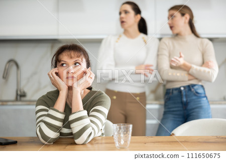 Upset middle-aged woman sitting at the kitchen table with her back to women quarreling to her Upset middle-aged woman sitting at the kitchen table with her back to women quarreling to her 111650675
