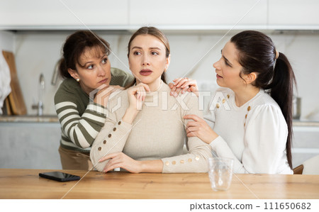 Sad middle-aged woman sitting at table while two others trying to calm her Sad middle-aged woman sitting at table while two others trying to calm her 111650682