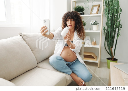 Pregnant woman blogger with a jar of pills and vitamins sits on the couch at home and takes pictures of herself on the phone, maintaining pregnancy and replenishing vitamins and minerals Pregnant woman blogger with a jar of pills and vitamins sits on the couch at home and takes pictures of herself on the phone, maintaining pregnancy and replenishing vitamins and minerals 111652519