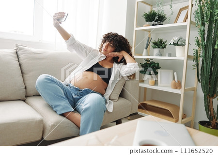 Pregnant woman blogger sits on the couch at home and takes pictures of herself on the phone, selfie and video call, consultation with the doctor online, pregnancy management 111652520