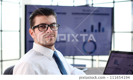 A businessman man in close-up in a shirt with graphs on the background looks at the camera A businessman man in close-up in a shirt with graphs on the background looks at the camera 111654194