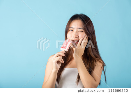 Portrait of Asian young woman with sensitive teeth after eating delicious ice cream wood stick mixed fruit flavor feeling painful uncomfortable, studio shot isolated on blue background, dental problem 111666250