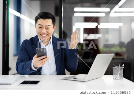 Happy Asian executive engaging in a virtual greeting, waving at the camera while having a video call on his mobile phone. 111666925