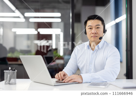 Focused Asian man in smart casual wear using laptop at desk with headset in a contemporary office setting. 111666946
