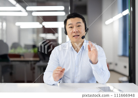 Asian businessman with headset in modern office providing customer support or leading a team meeting, displaying engagement. 111666948