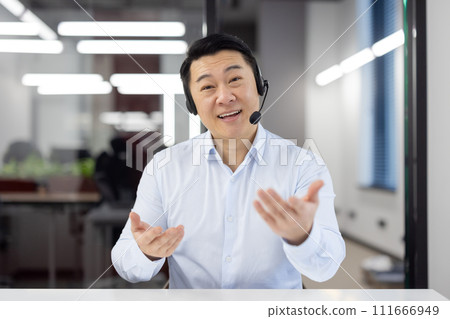 A cheerful Asian businessman in a headset actively engaging in conversation in an office setting. A cheerful Asian businessman in a headset actively engaging in conversation in an office setting. 111666949