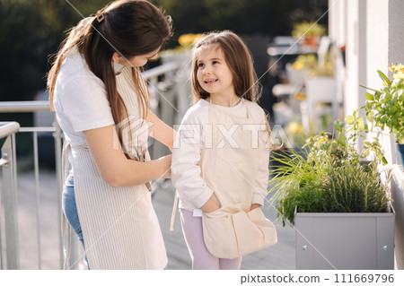 Beautiful young mom helps her daughter to get dressed overall for gardening on balcony. Adorable girl smile 111669796