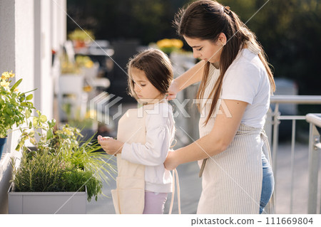 Beautiful young mom helps her daughter to get dressed overall for gardening on balcony. Adorable girl smile 111669804