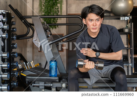 A young Asian man doing muscle training while sitting on a training bench at the gym (arm training, dumbbells, dumbbell curls) A young Asian man doing muscle training while sitting on a training bench at the gym (arm training, dumbbells, dumbbell curls) 111669960