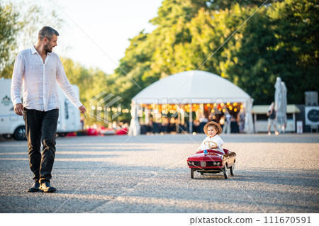 A smiling father rides a happy child in a red toy car A smiling father rides a happy child in a red toy car 111670591