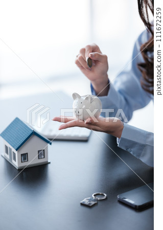 Close up Asian women's hand saving a coin into piggy bank with wooden house model on the table for business, finance, saving money and property investment concept. Close up Asian women's hand saving a coin into piggy bank with wooden house model on the table for business, finance, saving money and property investment concept. 111672259