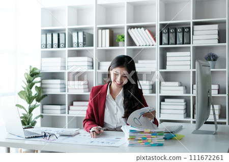 Beautiful asian businesswoman working on laptop and calculating with financial calculator from statistics data graphs, charts. Successful business results in modern office wear red shirt. 111672261
