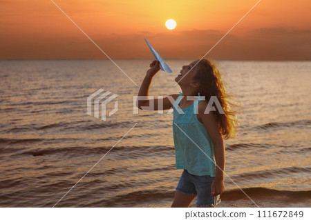 Little girl with paper plane in her hands playing on beach by sea o sunset Little girl with paper plane in her hands playing on beach by sea o sunset 111672849