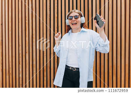 Closeup portrait of a dancing young woman listening to music through wireless earphones on wooden rustic background. Music lover enjoying music. Closeup portrait of a dancing young woman listening to music through wireless earphones on wooden rustic background. Music lover enjoying music. 111672959