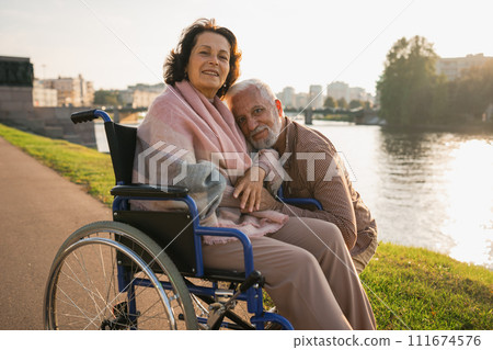 Senior woman in wheelchair walking with caregiver old man on road. Elderly family couple man supporting taking care of paralyzed woman in chair for people with disability outdoor. Rehabilitation Senior woman in wheelchair walking with caregiver old man on road. Elderly family couple man supporting taking care of paralyzed woman in chair for people with disability outdoor. Rehabilitation 111674576