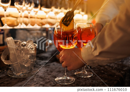 Bartender makes two glasses of cocktail Aperol spritz on bar counter, adds fresh orange slices. Typical alcoholic Italian beverage, aperitif made with Prosecco sparkling white wine and ice cubes. 111676010