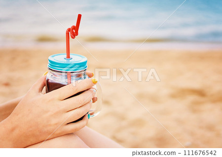 Glass jar with a drink and a straw in women's hands. Female sitting on the sea beach with an alcoholic cocktail. Vacation on tropical coast. Photo card with copy space. 111676542