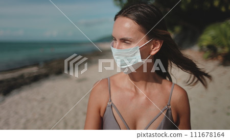 Woman walking summer beach wearing medical mask. Young female in bikini enjoy outdoor lifestyle travel on holiday vacation. Quarantine and self-isolation, epidemic pandemic 111678164
