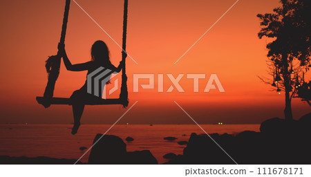 Happy woman silhouette swinging in tropical sunset sitting on swing with scenic view on orange sky, sea waves. Outdoor lifestyle travel on summer holiday vacation, enjoy freedom, have fun. Happy woman silhouette swinging in tropical sunset sitting on swing with scenic view on orange sky, sea waves. Outdoor lifestyle travel on summer holiday vacation, enjoy freedom, have fun. 111678171