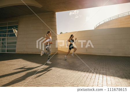 Two young women in sportswear are running on modern buildings background. Active lifestyle concept Two young women in sportswear are running on modern buildings background. Active lifestyle concept 111678615