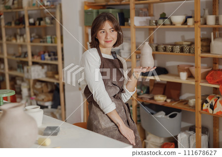 Portrait of young female potter in apron with mug looks away while posing in workshop 111678627
